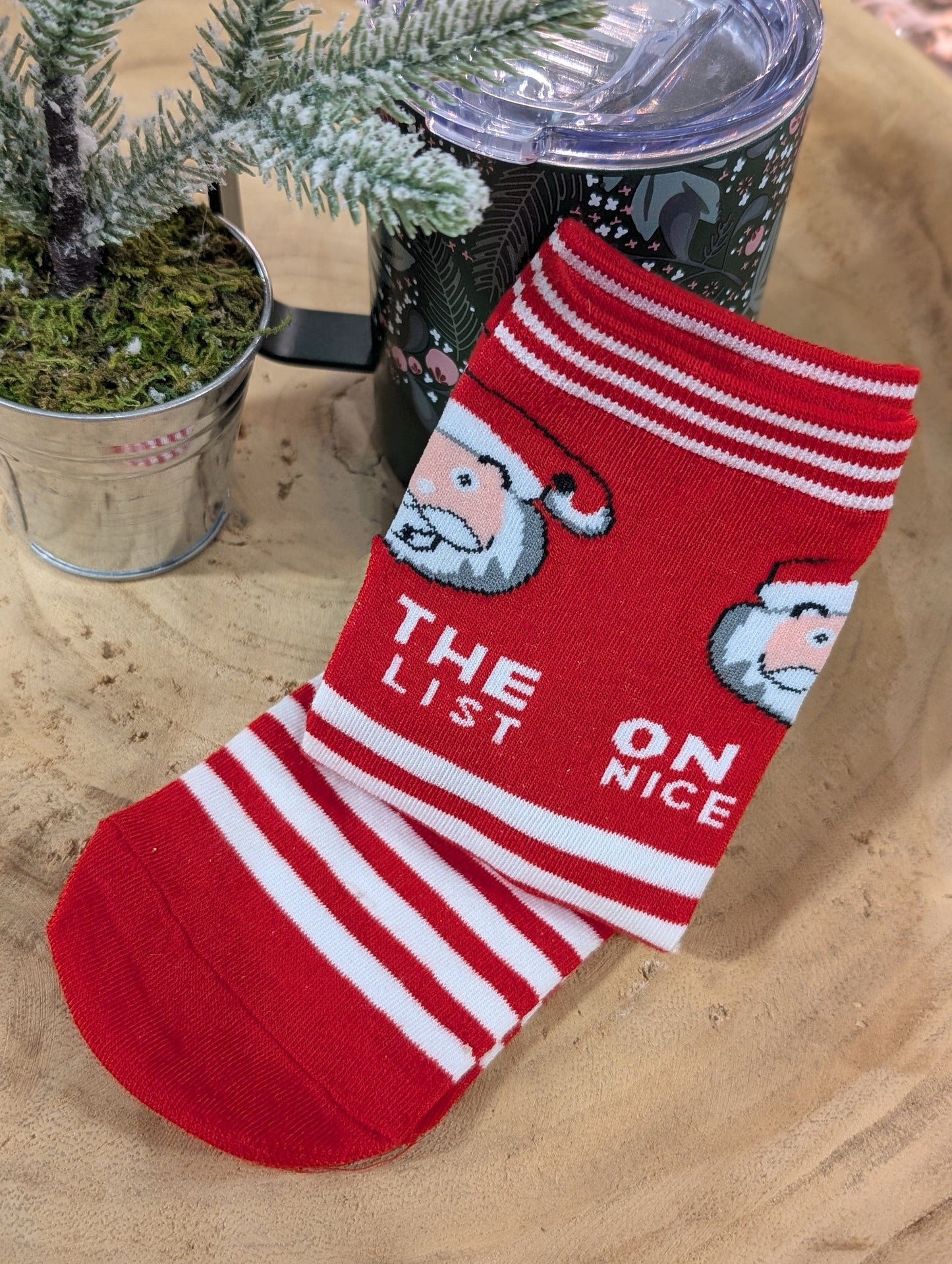Cozy red and white holiday socks featuring Santa faces and stripes, with "On the nice list" written across them. They're elegantly displayed on a wooden surface beside a small potted plant, capturing the festive spirit.