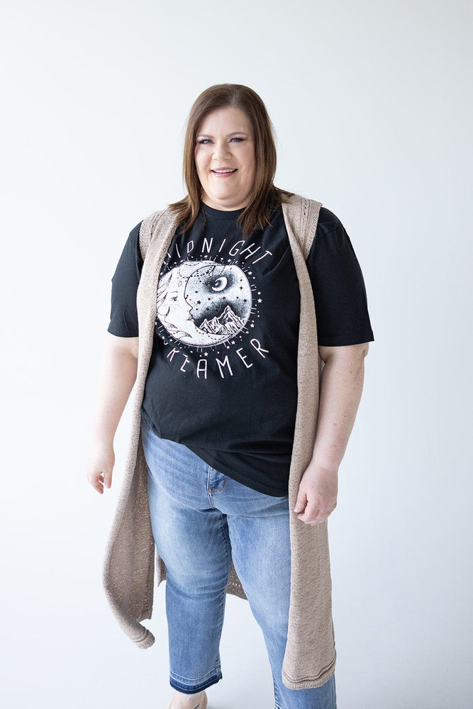 A person with long brown hair, wearing a black "Midnight Dreamer" t-shirt, light blue jeans, and a versatile beige OPEN WEAVE DUSTER VEST by Yesta, stands and smiles at the camera against a plain white background.