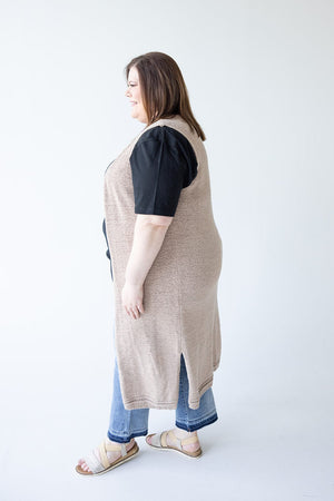 A person is standing sideways, showcasing a versatile look with a black t-shirt, the Yesta OPEN WEAVE DUSTER VEST in beige, blue jeans, and sandals. Perfect for a mid to plus-size wardrobe, the plain white background emphasizes their stylish yet effortless attire.