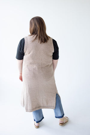 A person with long hair is standing with their back to the camera, wearing the Yesta OPEN WEAVE DUSTER VEST over a black top and blue jeans, paired with beige sandals. The vest's luxurious fabric adds a touch of elegance to this mid to plus-size wardrobe staple.