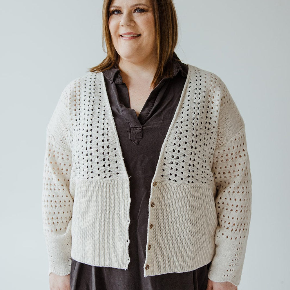 A person with shoulder-length hair smiles while wearing a dark dress and the Love Marlow OPEN WEAVE KNIT CARDIGAN, featuring wooden buttons. The oversized relaxed fit of the cardigan adds a cozy touch as they stand against a plain backdrop.