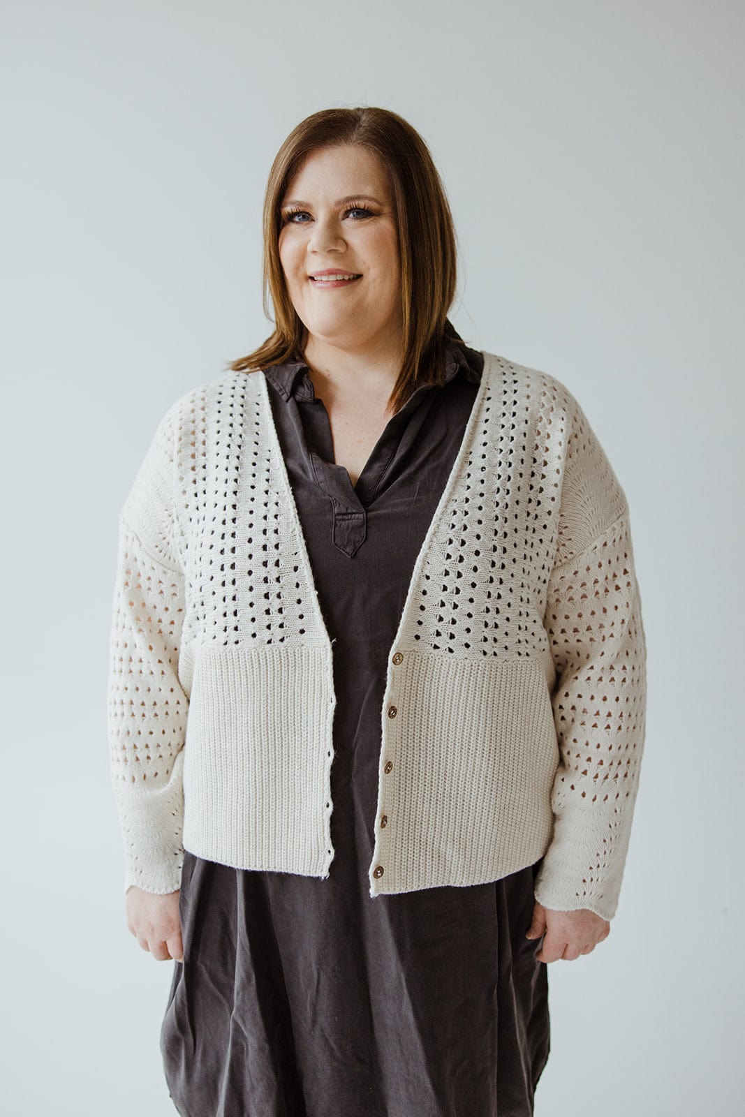 A person with shoulder-length hair smiles while wearing a dark dress and the Love Marlow OPEN WEAVE KNIT CARDIGAN, featuring wooden buttons. The oversized relaxed fit of the cardigan adds a cozy touch as they stand against a plain backdrop.