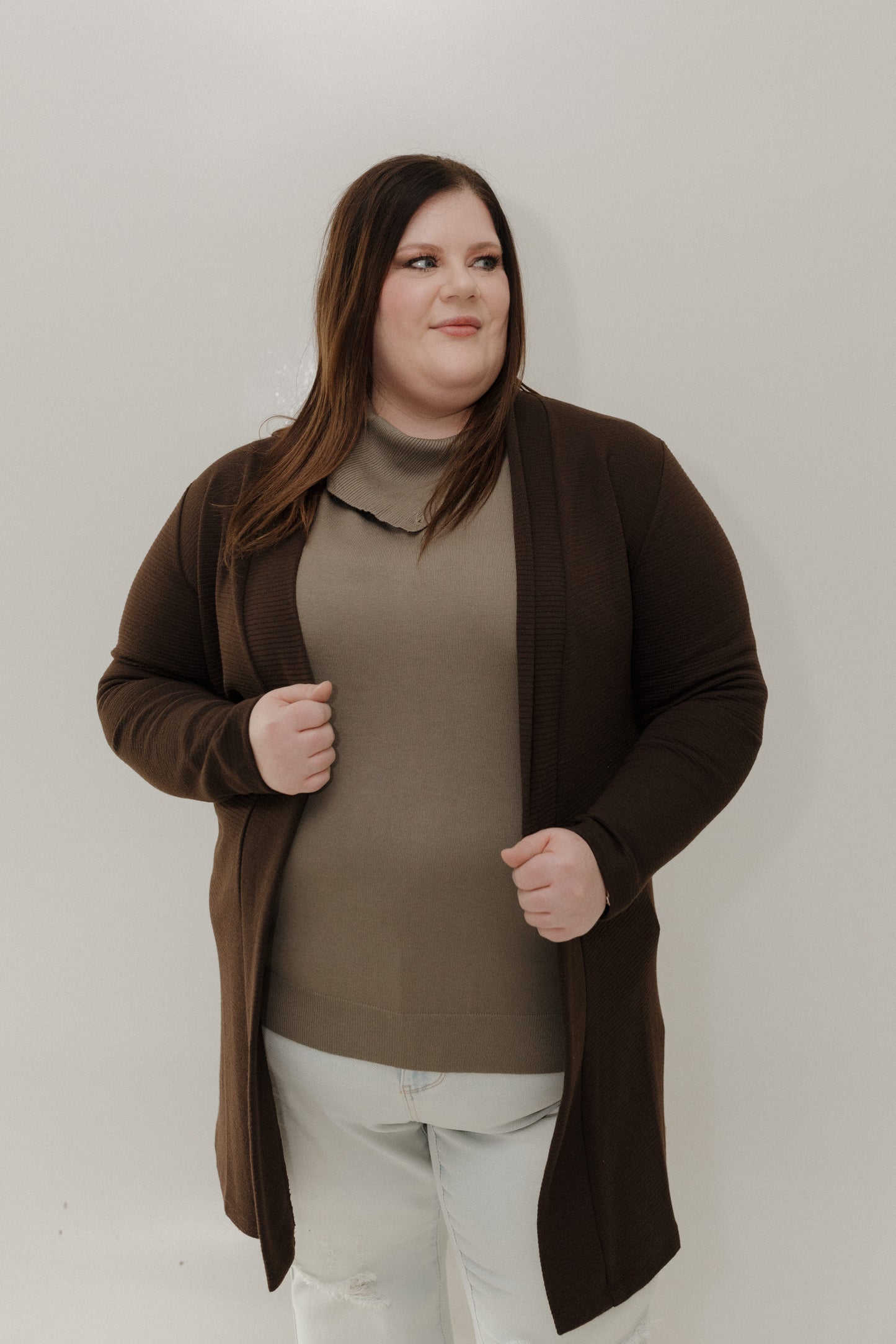Woman wearing a brown cardigan over a beige sweater and white pants against a plain background