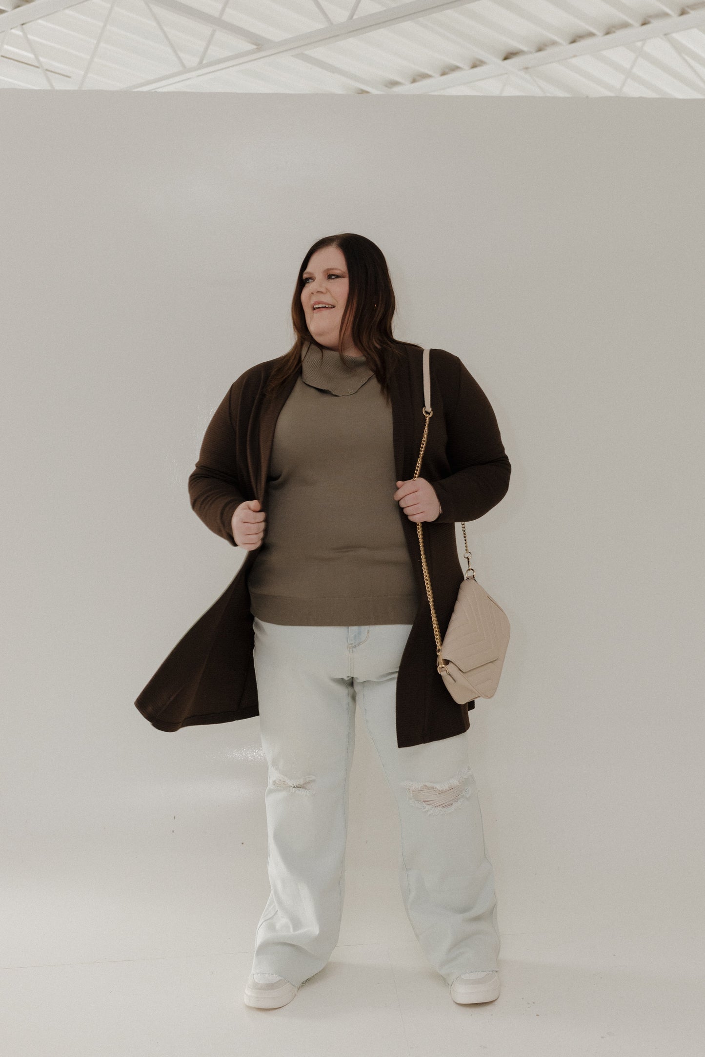 Person wearing a brown coat, green sweater, and light jeans holding a beige handbag against a plain background.
