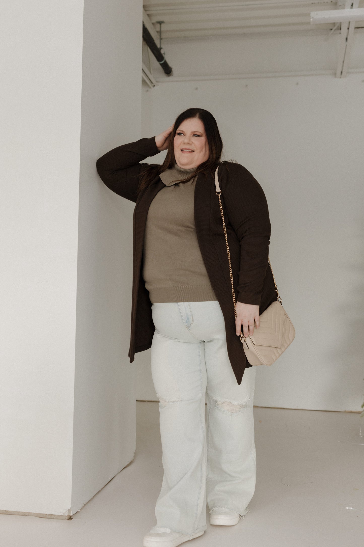 Person wearing a brown coat, beige sweater, and light blue jeans in an indoor setting.