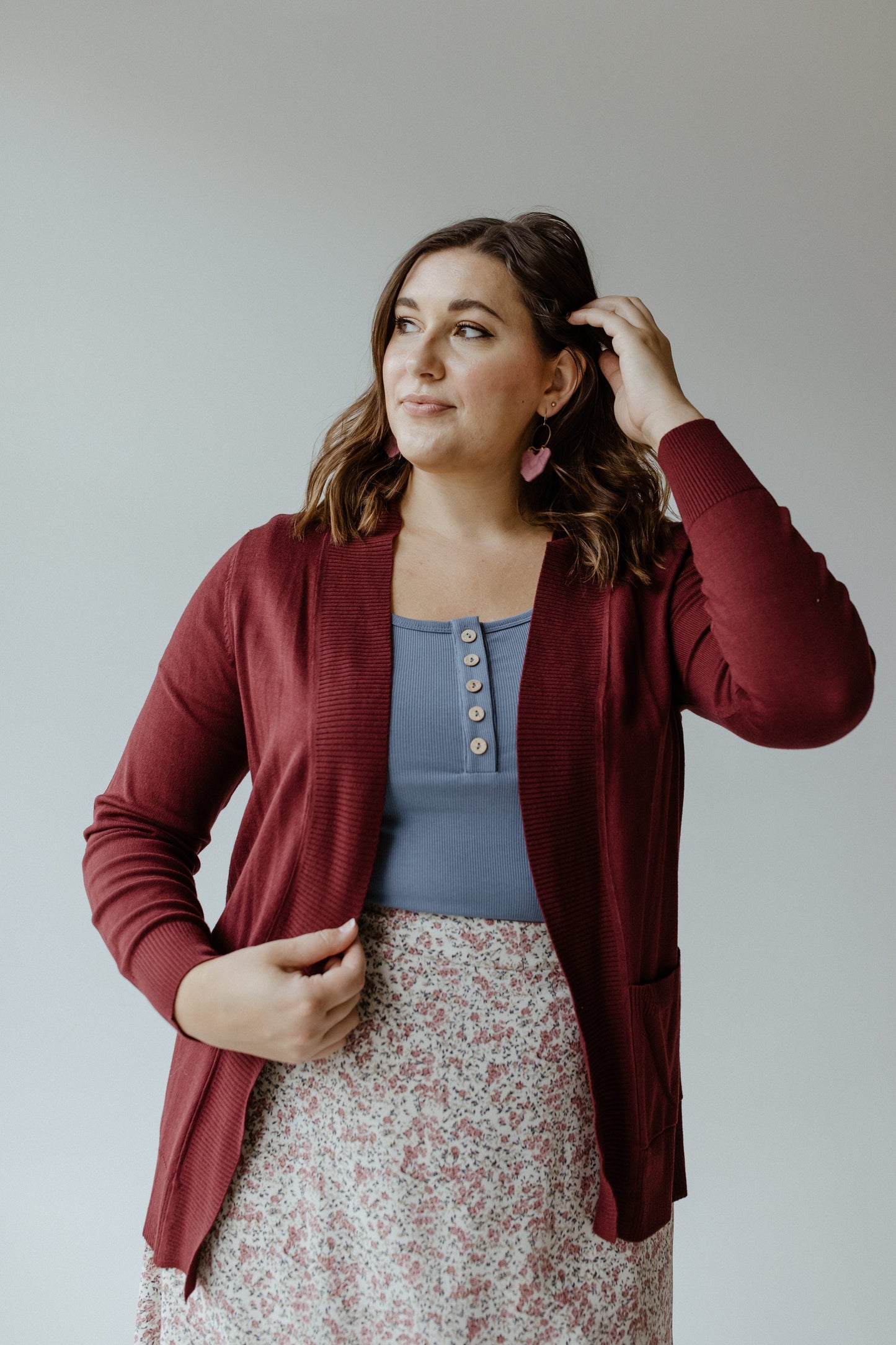 A woman stands against a plain background, exuding a cozy feel in her burgundy cardigan, which complements her blue top and floral skirt. The lightweight fabric drapes elegantly as she touches her hair with one hand.