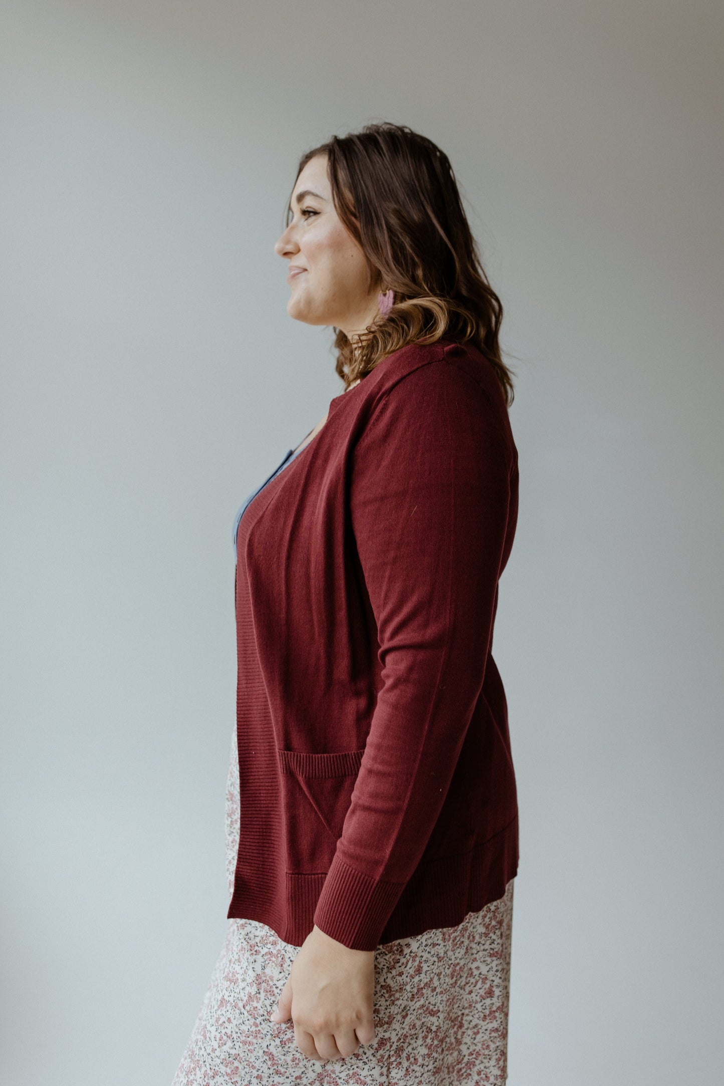 A woman in a side profile wears a burgundy cardigan with a cozy feel over a floral dress against a plain background.