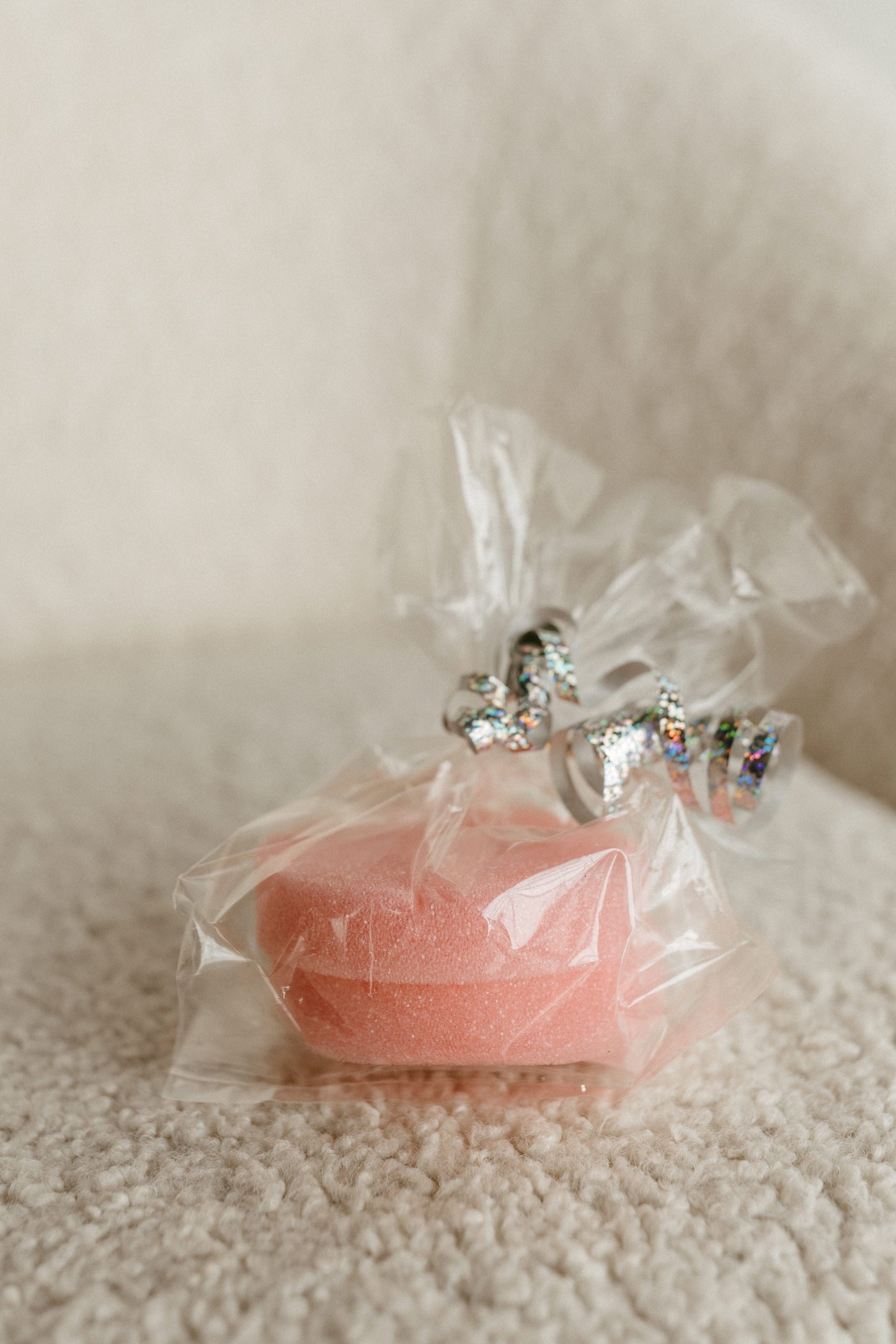 A transparent plastic wrap contains a pink, heart-shaped sponge, perfect for those wardrobe malfunctions. Tied with a decorative silver ribbon, it's elegantly placed on a textured light beige surface. Ideal as an OPPS ERASER DEODORANT REMOVER to tackle unwanted deodorant stains.