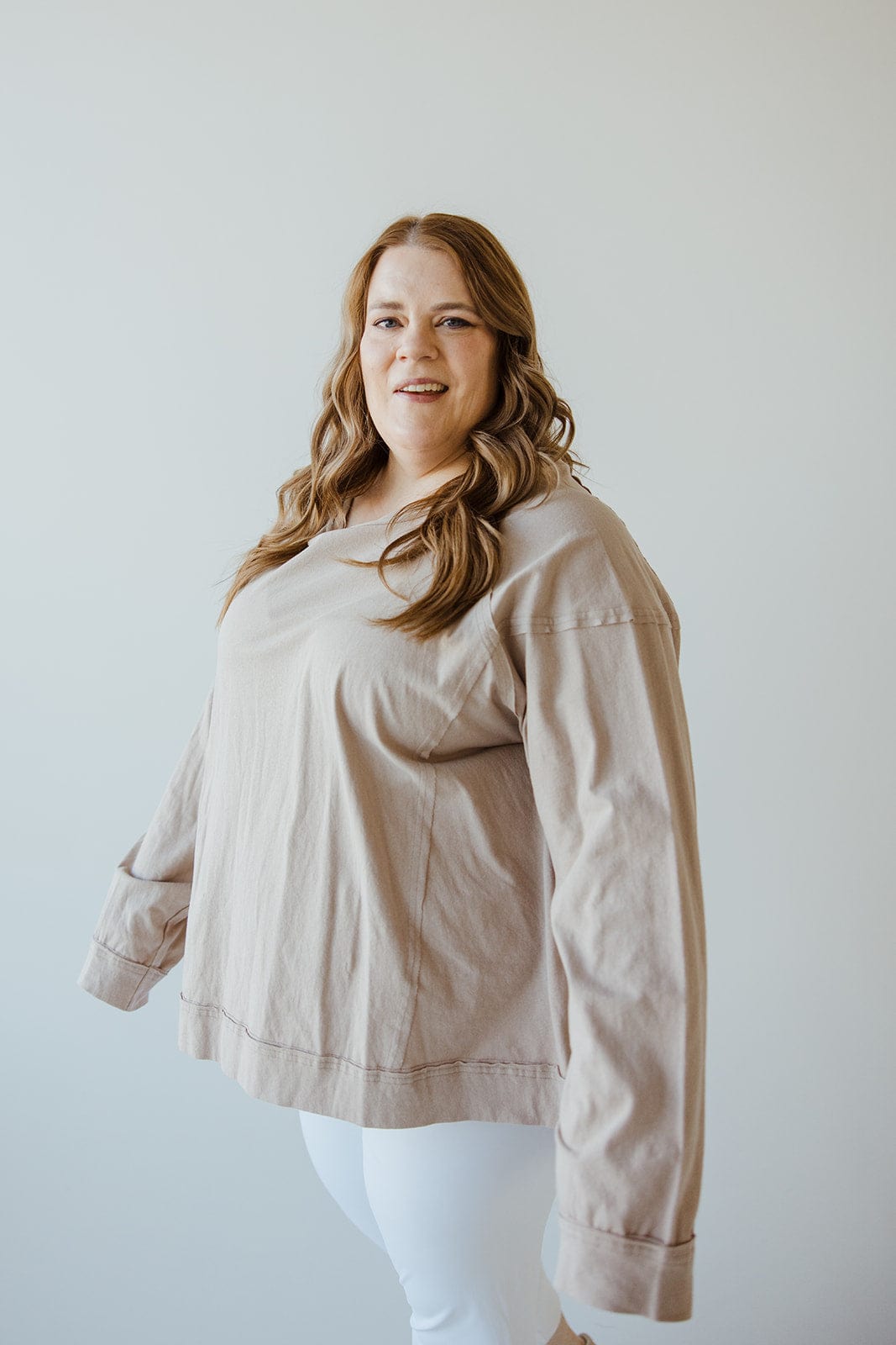 A person with long hair, wearing a lightweight knit beige top and white pants, stands against a plain white background.