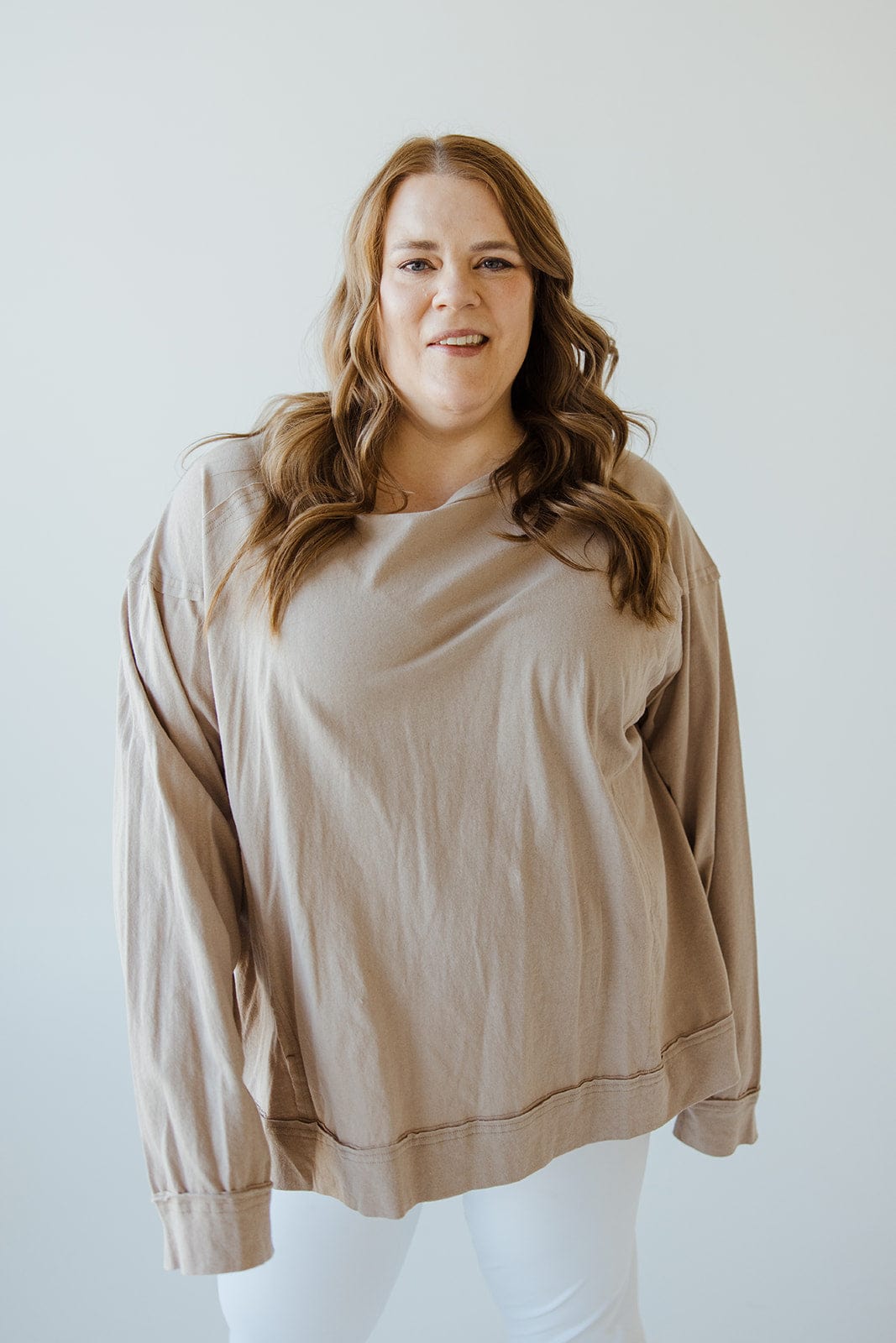 A person with long hair wearing an oversized long sleeve tee featuring raw seam detail and white pants stands against a plain background.