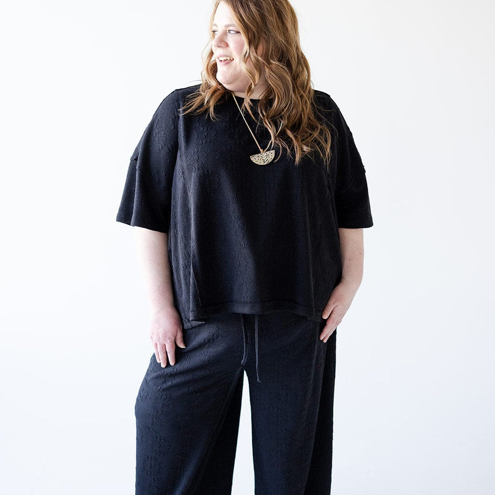 A person with long hair is wearing the Love Marlow Oversized Subtle Floral Pattern Dolman Tee and sandals, standing with an athleisure vibe against a plain white background, glancing to the side.