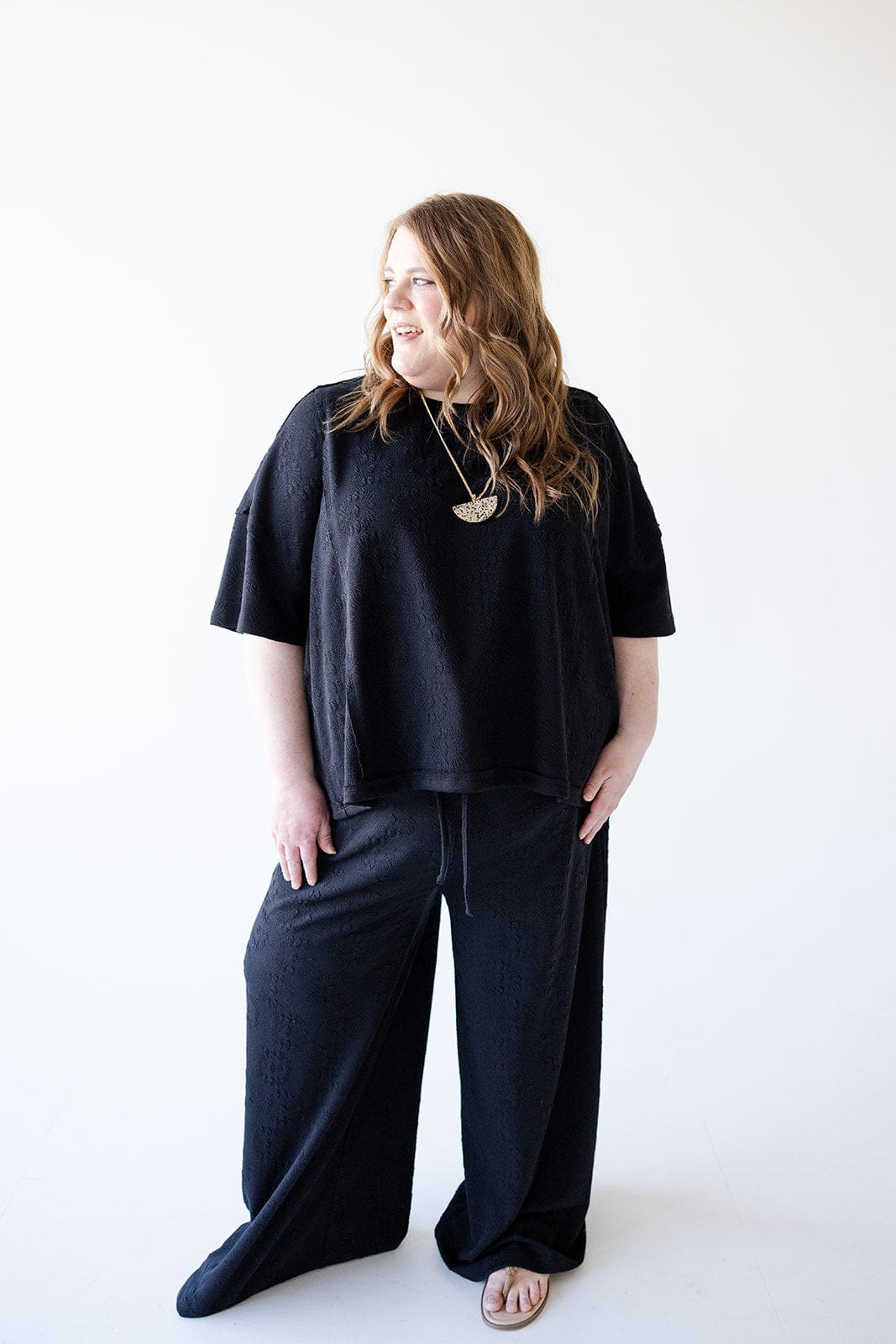 A person with long hair is wearing the Love Marlow Oversized Subtle Floral Pattern Dolman Tee and sandals, standing with an athleisure vibe against a plain white background, glancing to the side.