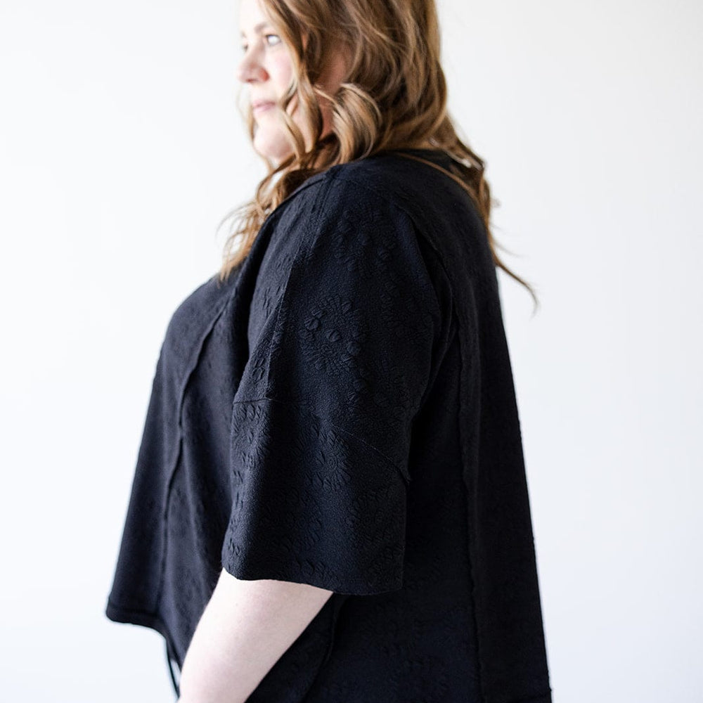 A person with long hair is wearing a black outfit with a subtle floral texture, the oversized Dolman tee from Love Marlow enhancing an athleisure vibe, standing in profile against a plain white background.