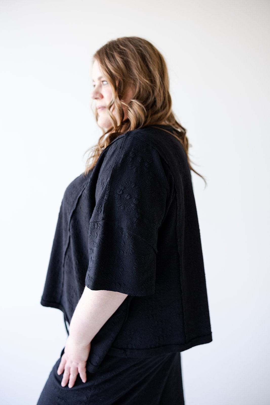 A person with long hair is wearing a black outfit with a subtle floral texture, the oversized Dolman tee from Love Marlow enhancing an athleisure vibe, standing in profile against a plain white background.