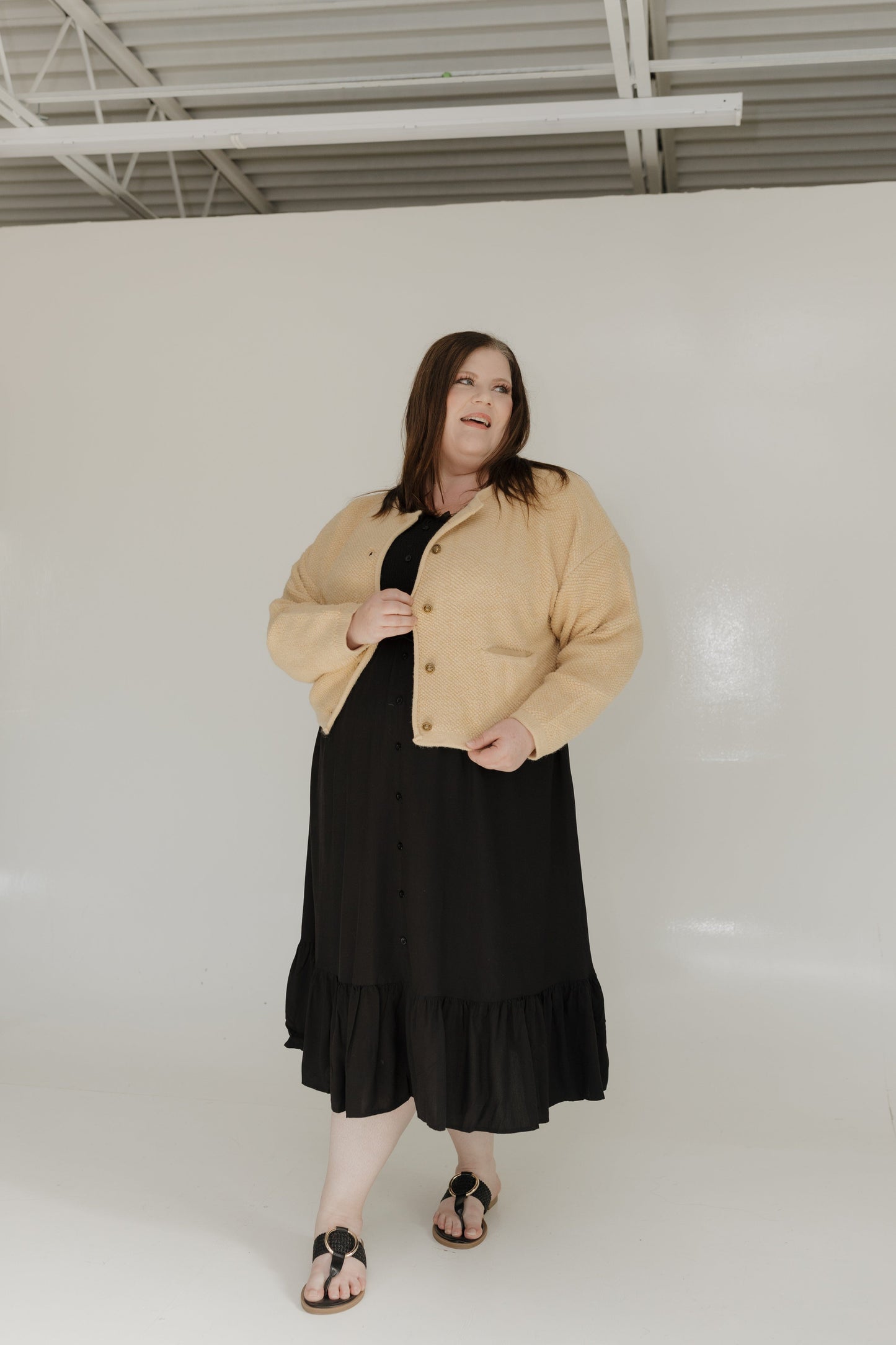 Woman wearing a beige jacket over a black dress in a white room.