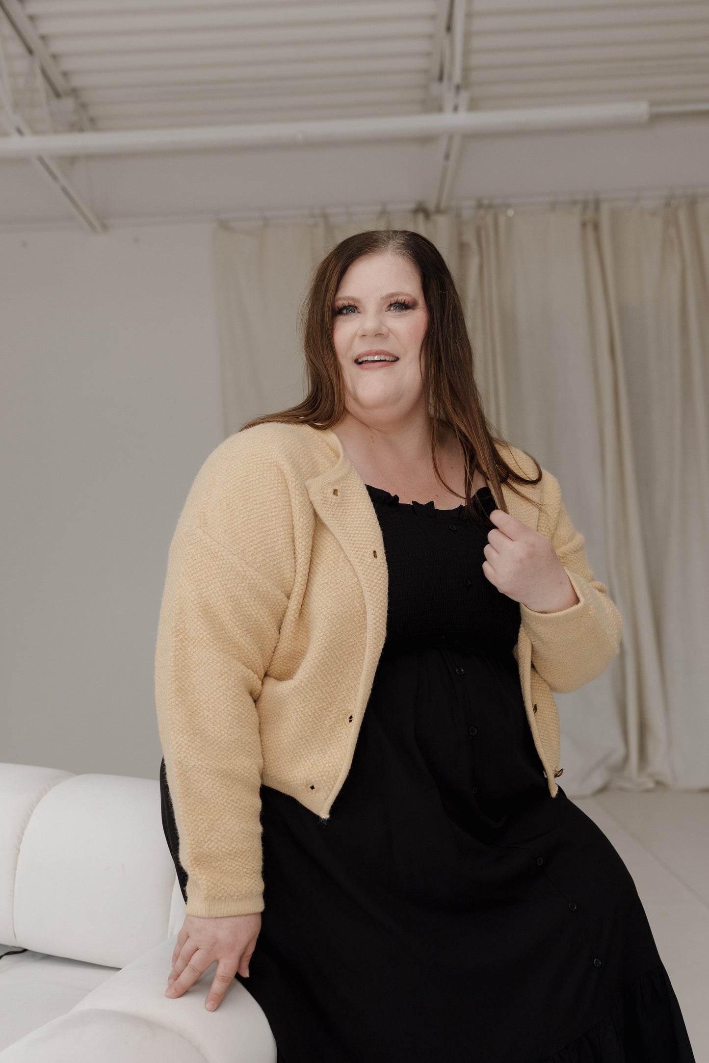 Woman wearing a beige cardigan over a black dress sitting on a white couch.