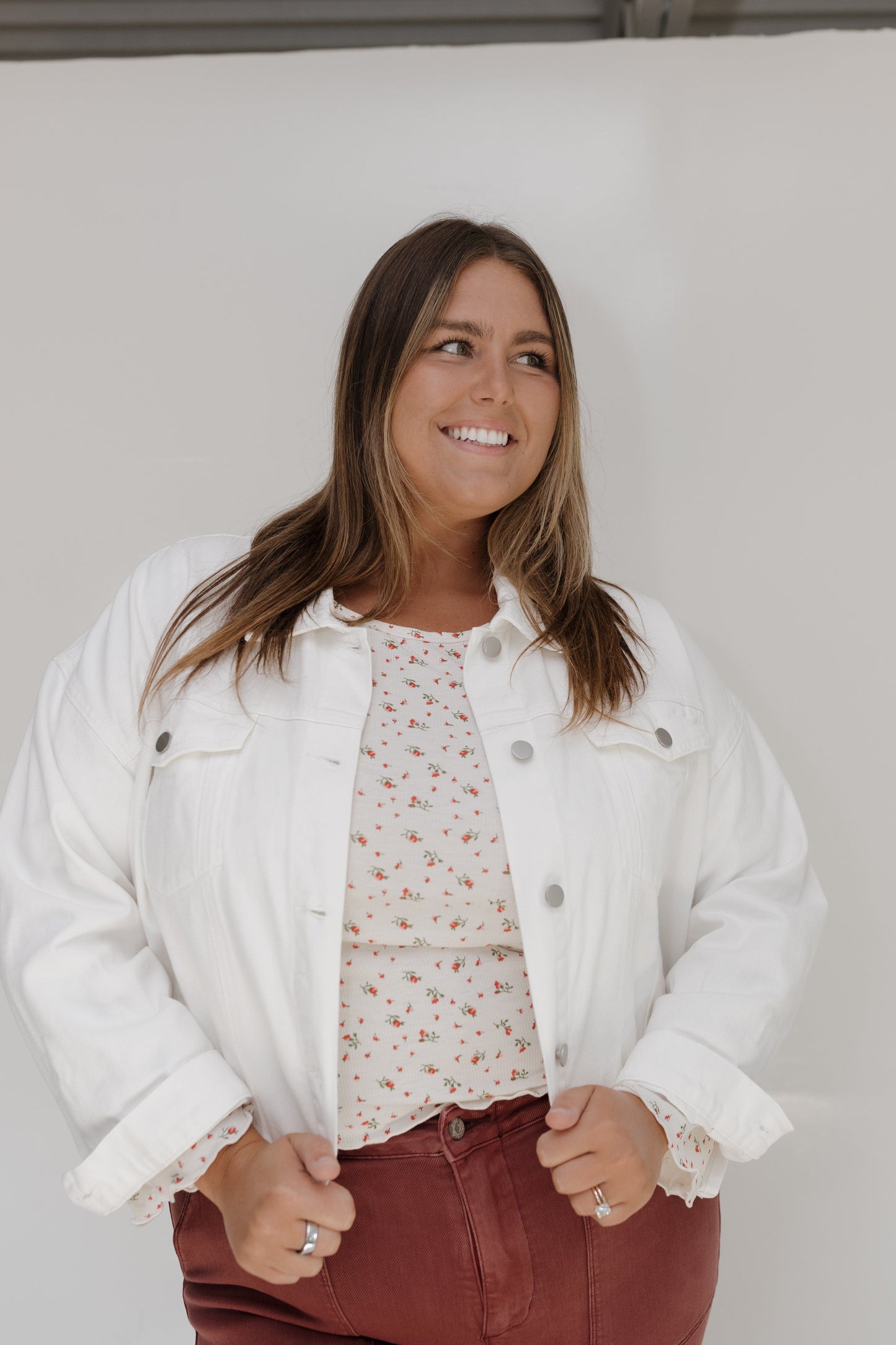 Woman wearing a white denim jacket over a floral top and red pants against a plain background