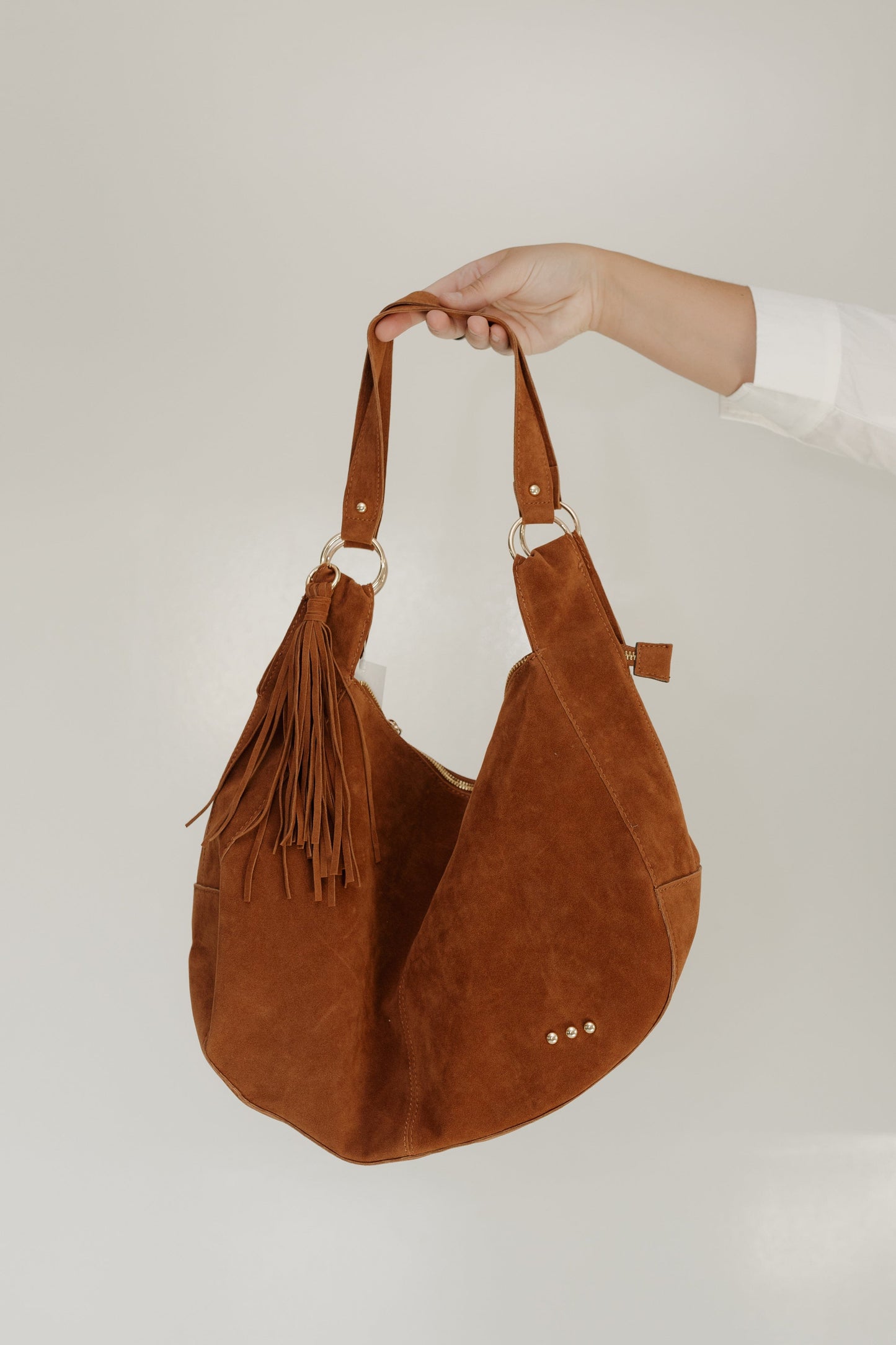 Brown suede handbag with tassels held by a person against a white background