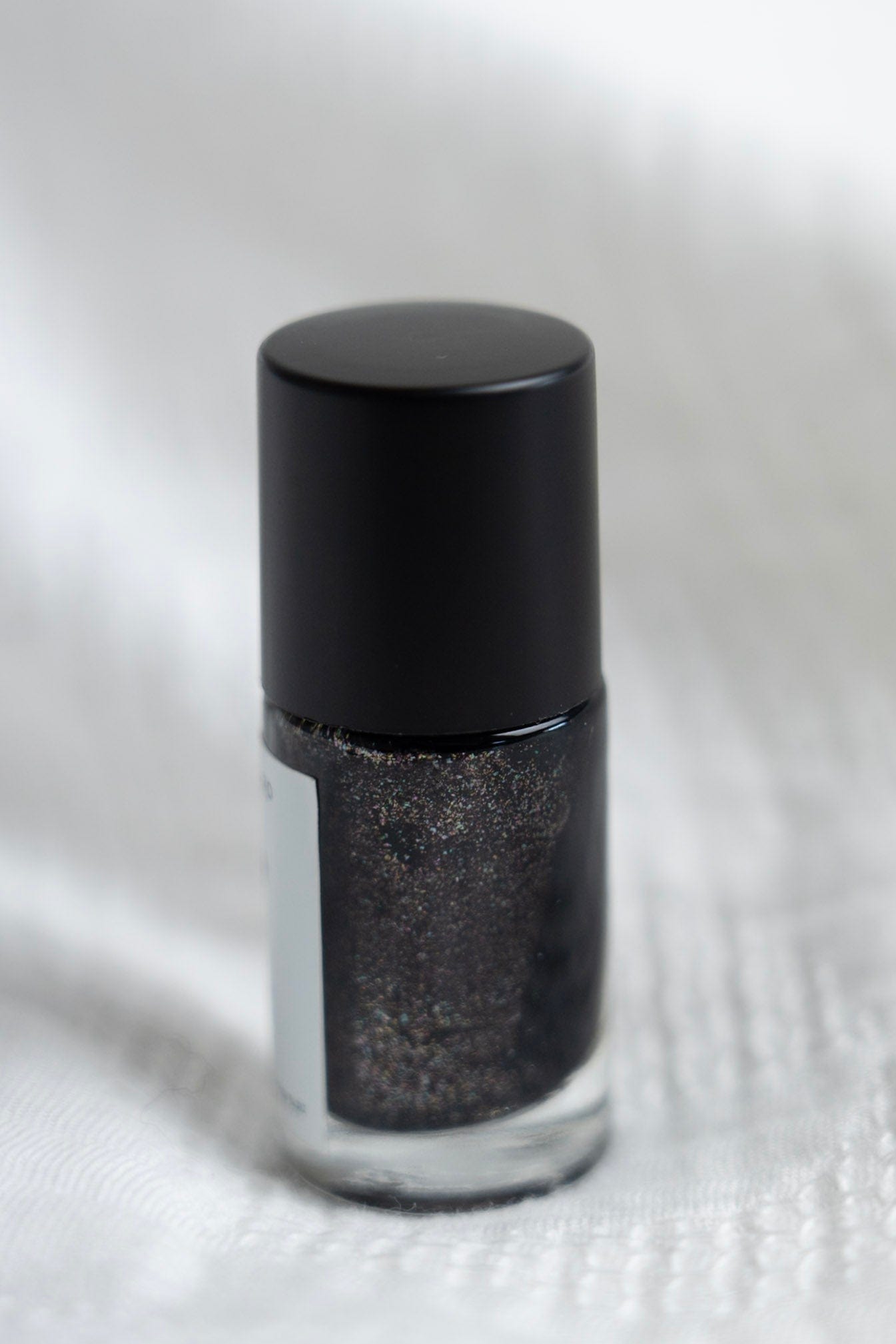 Black nail polish bottle with glittery black polish on a textured white background