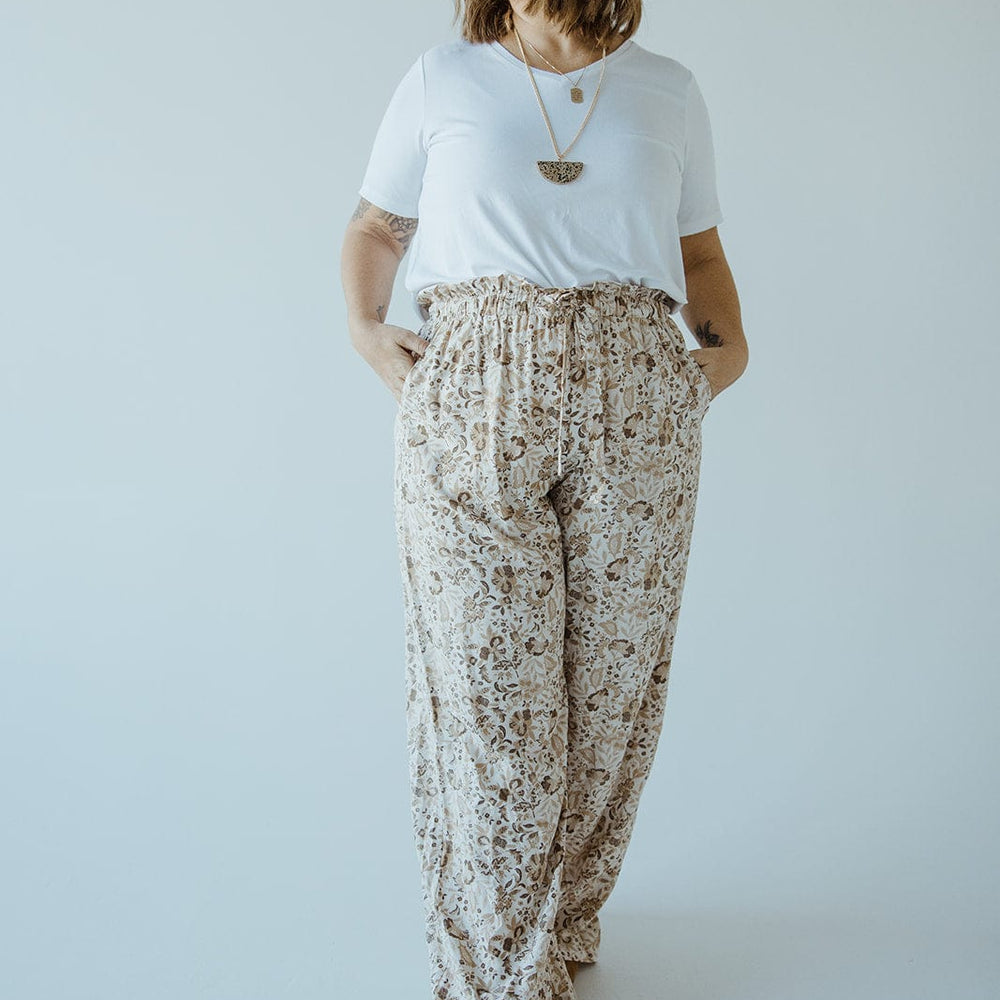 A woman stands against a plain background, wearing a white T-shirt, brown shoes, and Love Marlow's PAPERBAG WAIST WIDE LEG PRINTED PANTS. She has her hands in her pockets and is looking away from the camera.