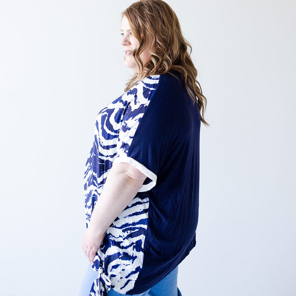 A woman with long, wavy hair wears a navy and white patterned PATTERN CHIFFON BLOUSE WITH KNIT BACK by Love Marlow with blue pants, standing against a plain white background, facing left with a slight smile.