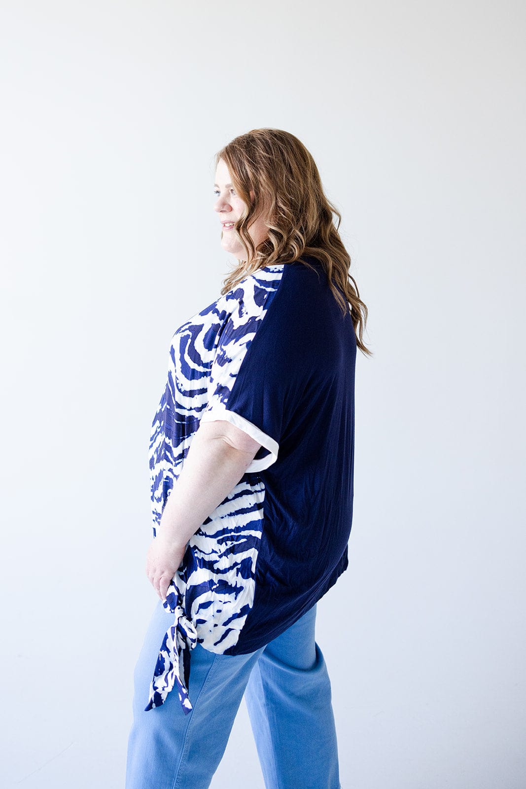 A woman with long, wavy hair wears a navy and white patterned PATTERN CHIFFON BLOUSE WITH KNIT BACK by Love Marlow with blue pants, standing against a plain white background, facing left with a slight smile.