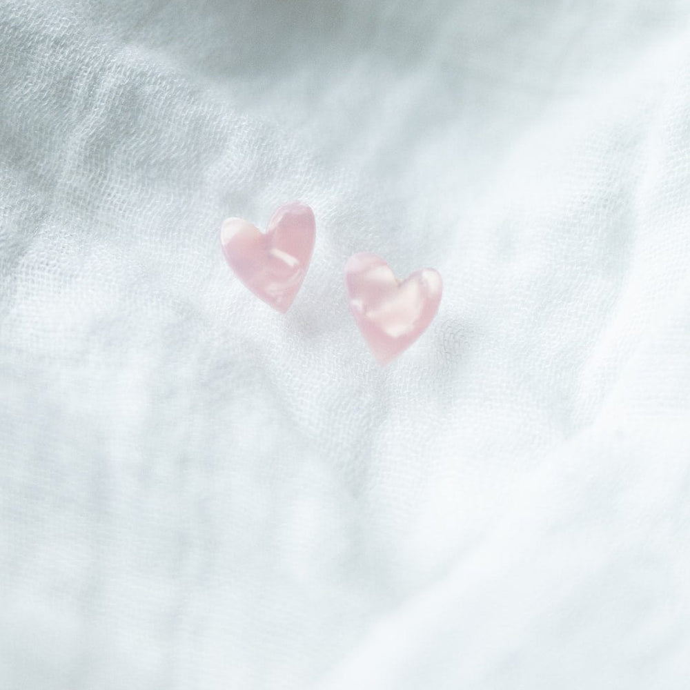 Two small pink heart studs rest elegantly on a textured white fabric background, crafted from plant-based acetate acrylic with hypoallergenic stainless steel posts for comfort and style.