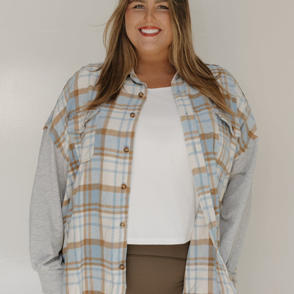 Woman wearing a plaid shirt over a gray long-sleeve shirt with a white top underneath, standing against a plain background.