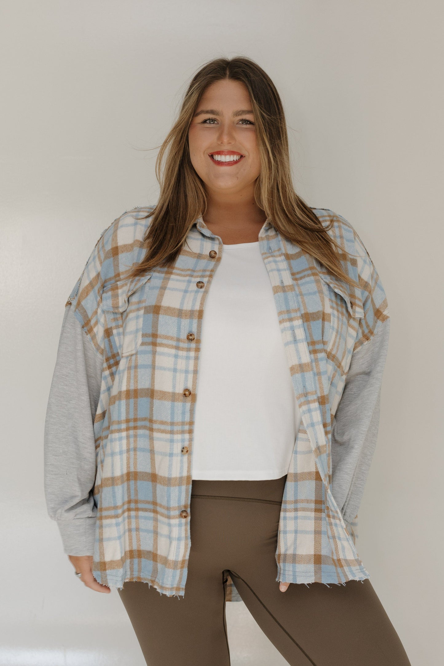 Woman wearing a plaid shirt over a gray long-sleeve shirt with a white top underneath, standing against a plain background.