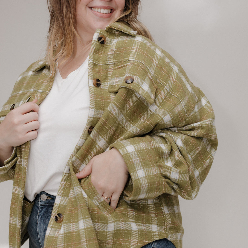 Woman wearing a green plaid jacket over a white shirt and blue jeans, standing against a plain background.