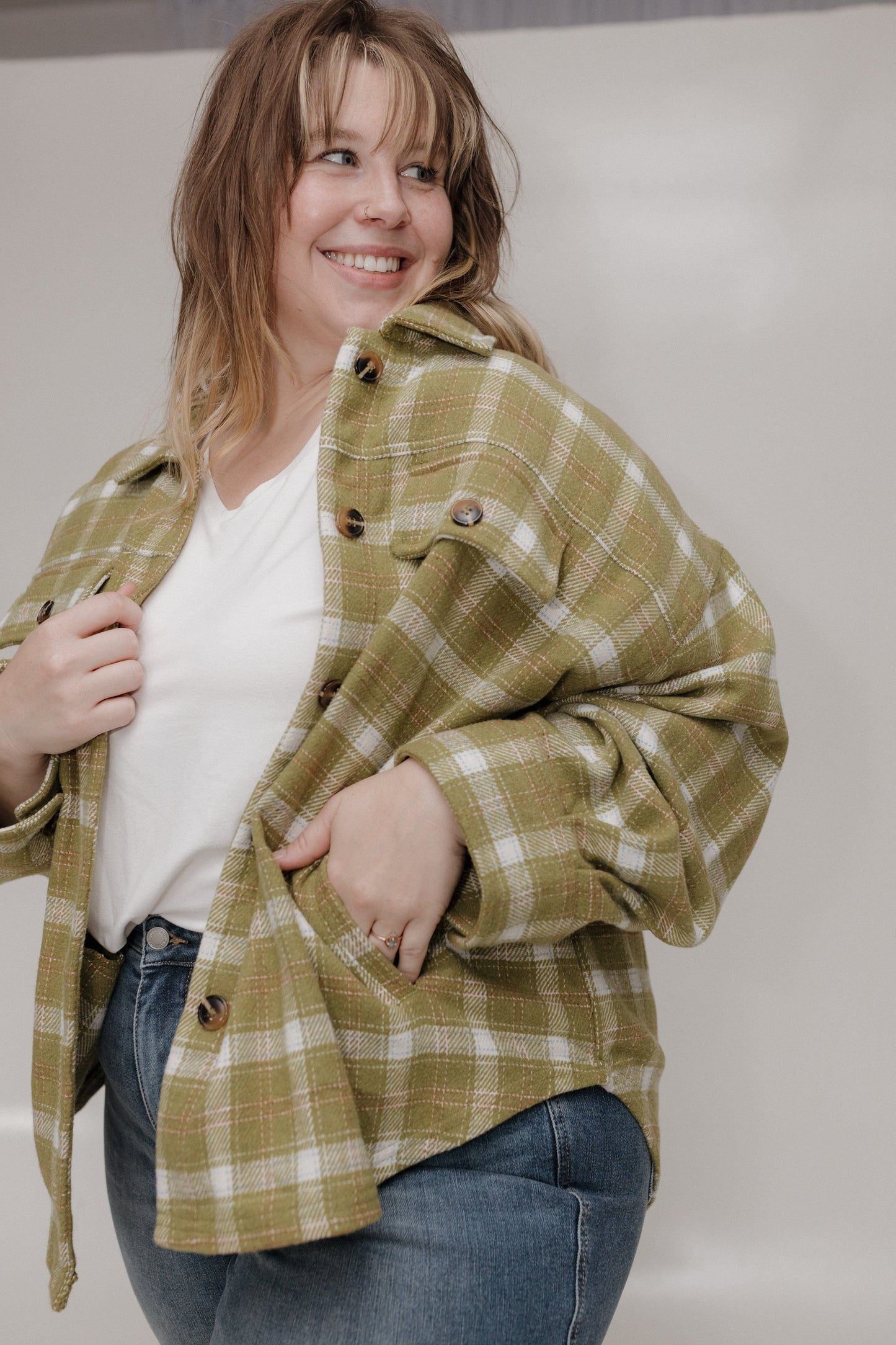 Woman wearing a green plaid jacket over a white shirt and blue jeans, standing against a plain background.