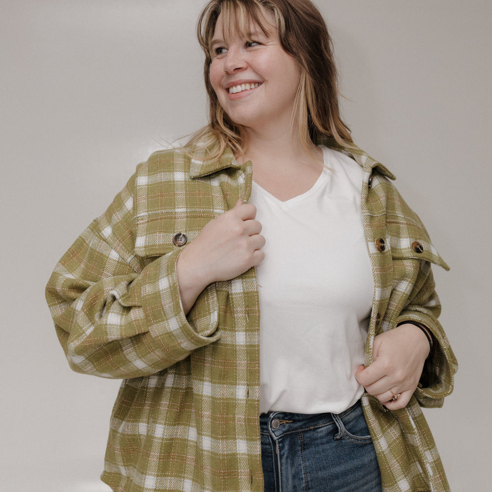 Woman wearing a green plaid shirt over a white top and blue jeans, standing against a plain background.