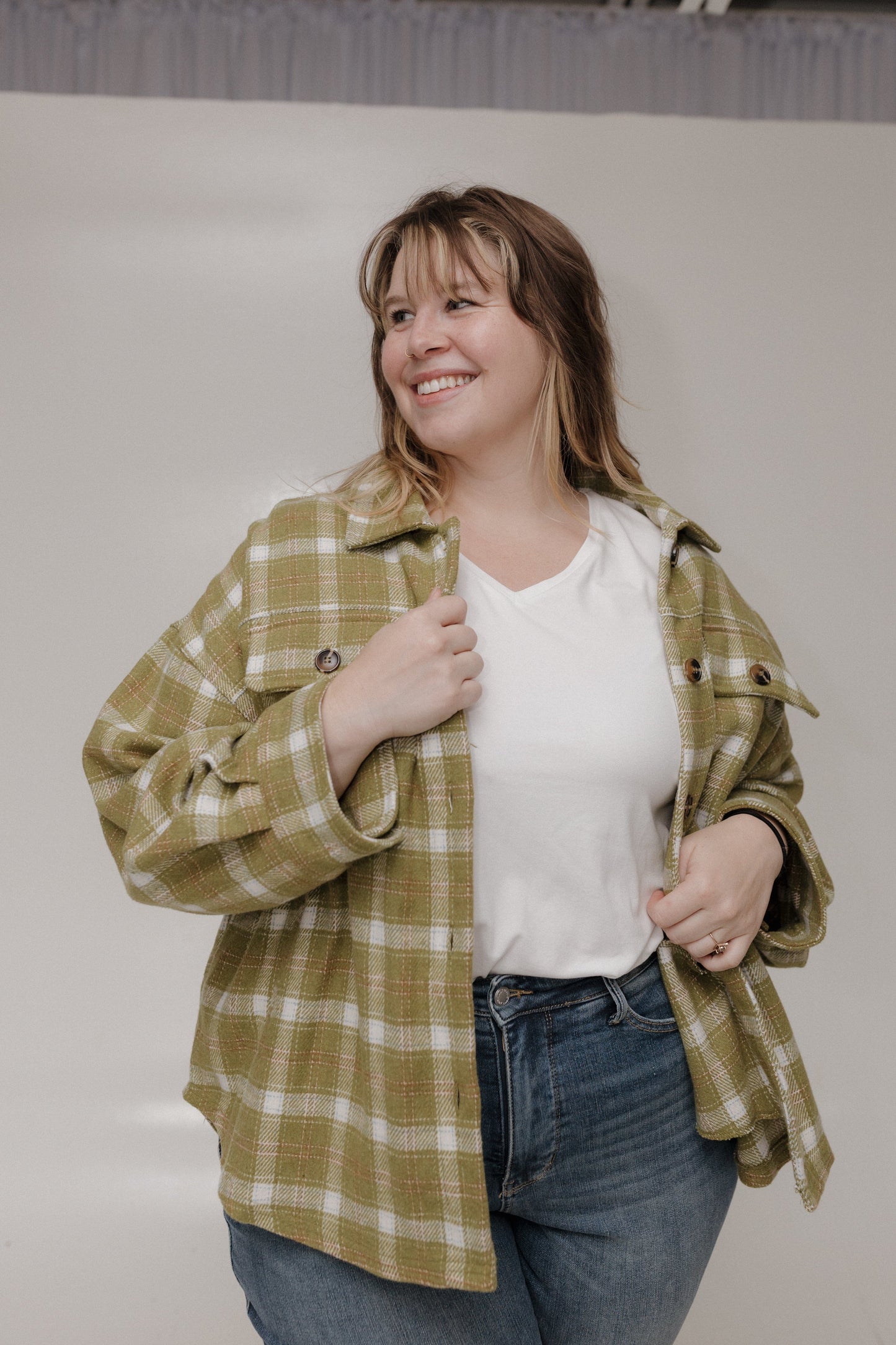Woman wearing a green plaid shirt over a white top and blue jeans, standing against a plain background.