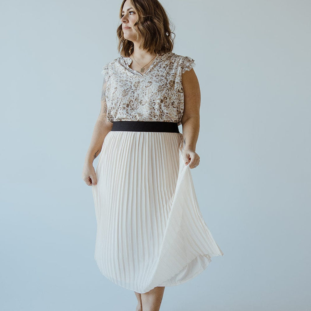 A woman stands in a light-colored room wearing a floral blouse and a taupe pleated midi skirt with an elasticated waist, gracefully holding the skirt with one hand.