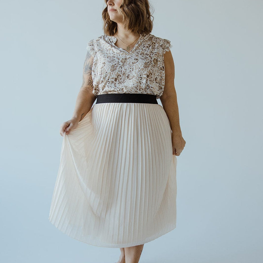 A woman with shoulder-length hair stands against a plain background, wearing a floral blouse and a taupe pleated midi skirt crafted from luxe chiffon fabric, featuring an elasticated waist for effortless elegance.