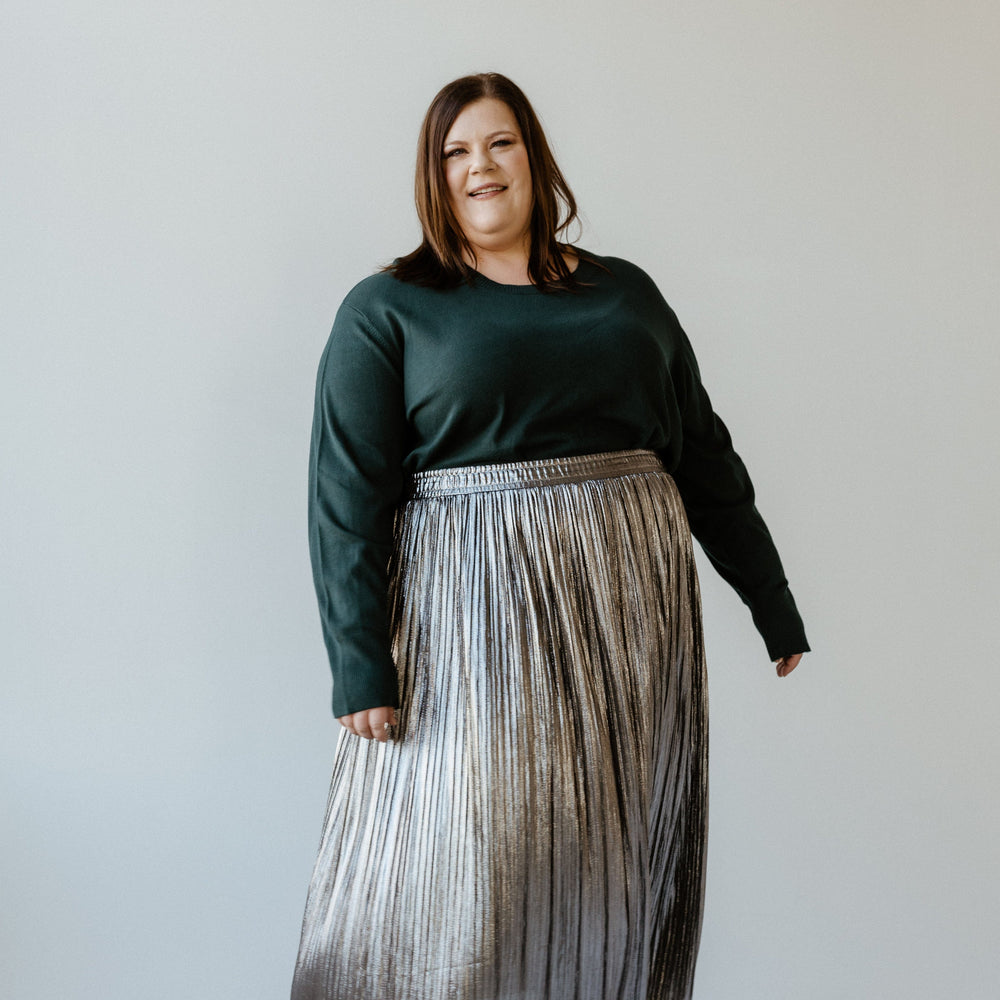 A woman in a dark sweater and a metallic silver pleated midi skirt stands against a plain background, smiling radiantly.