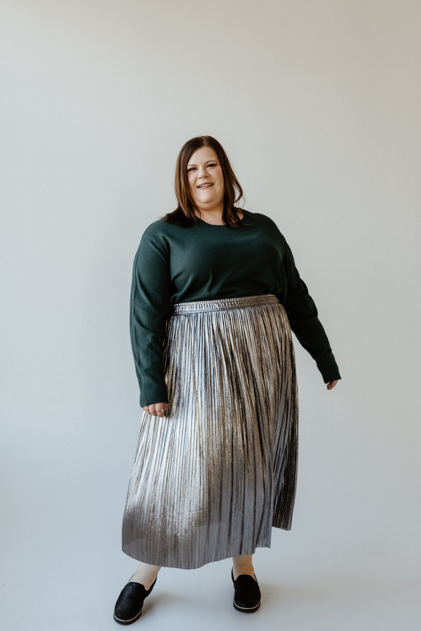 A woman in a dark sweater and a metallic silver pleated midi skirt stands against a plain background, smiling radiantly.