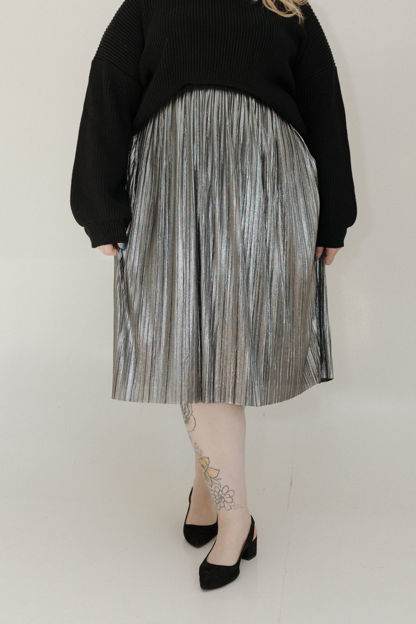 Person wearing a black sweater and silver pleated skirt on a plain background