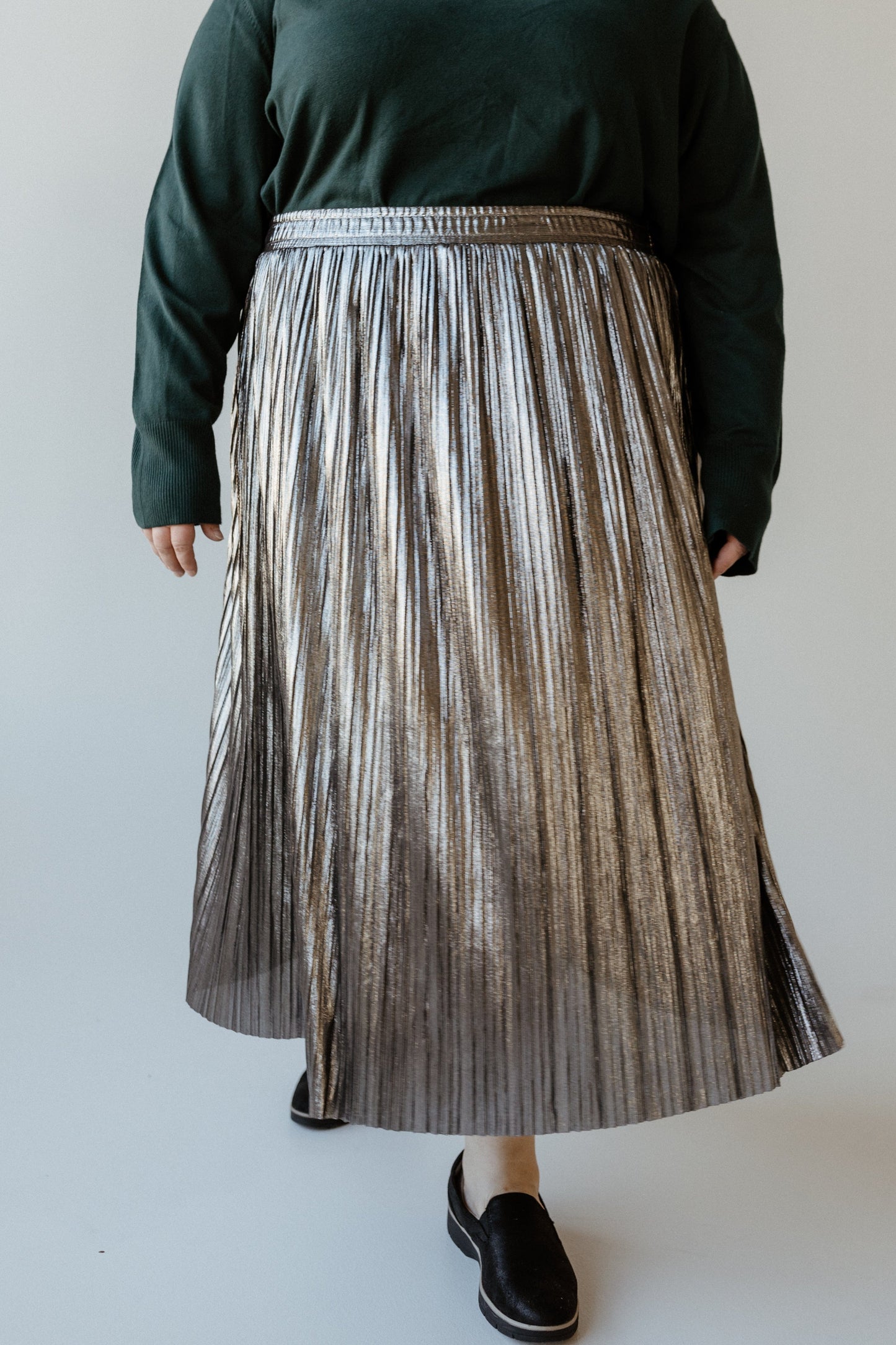 A person wearing a green sweater and a pleated midi skirt in metallic silver, paired with black shoes, stands confidently against a plain background.