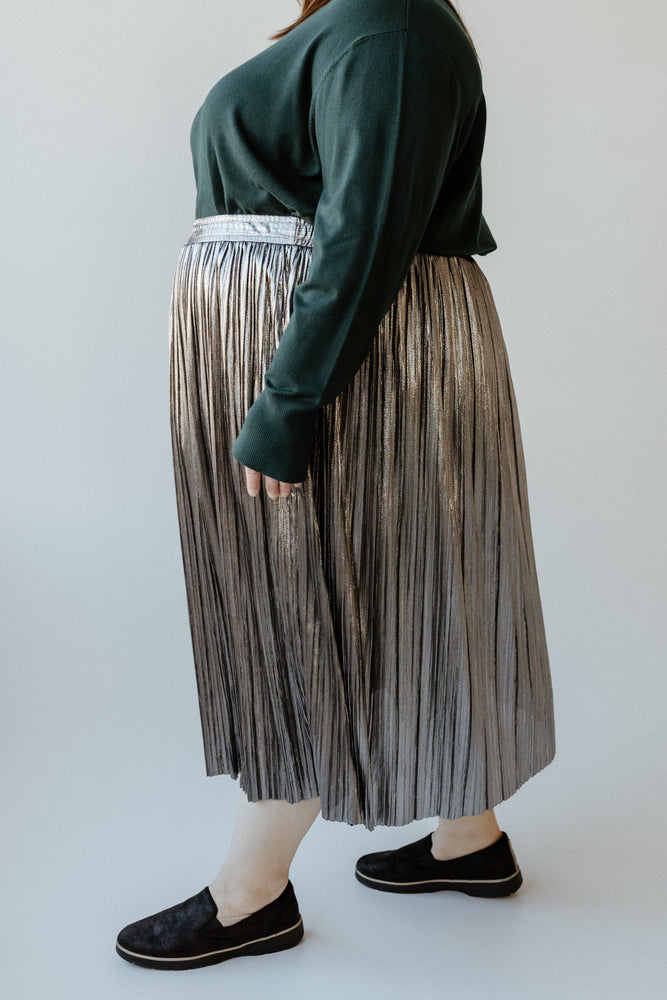 A person is wearing a green sweater, a metallic silver pleated midi skirt, and black slip-on shoes.