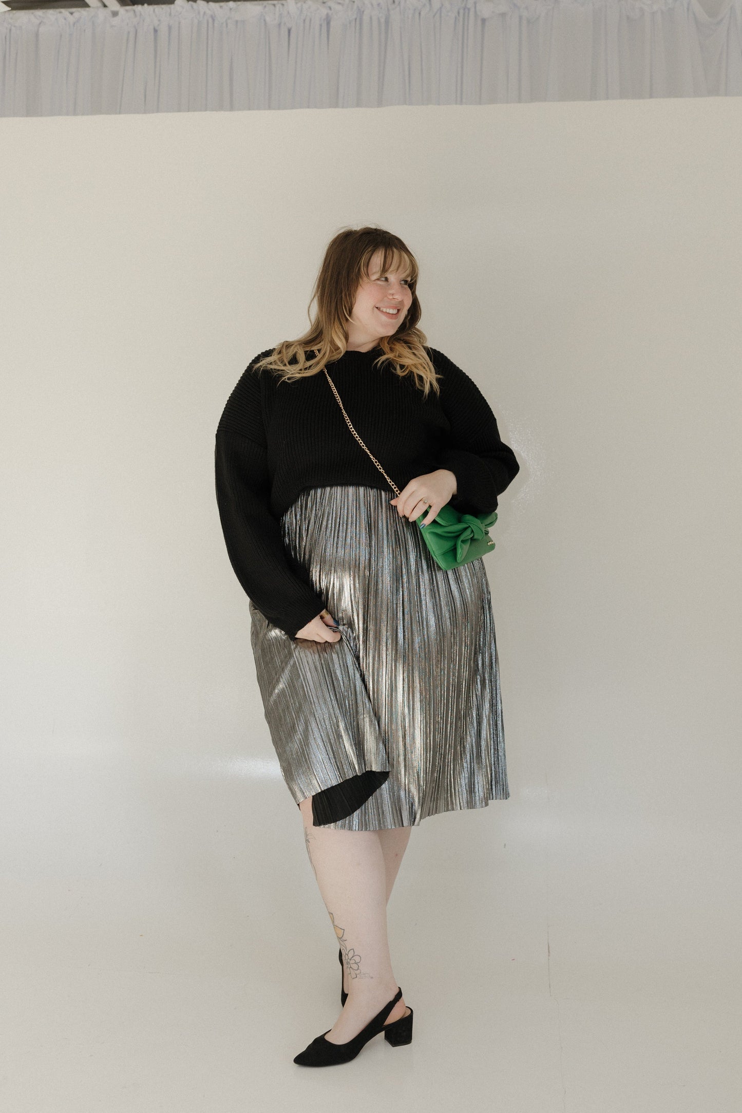 Woman wearing a black sweater and silver skirt holding a green clutch against a white background