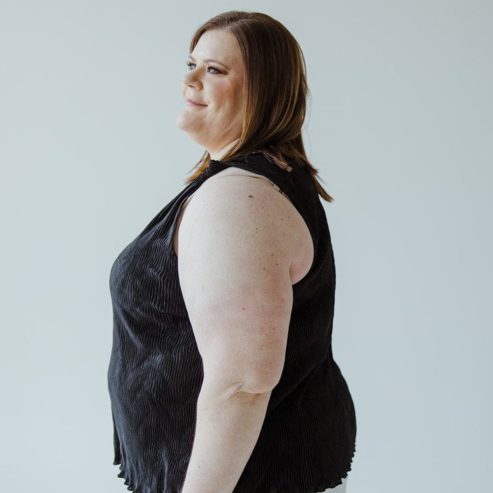 Person with shoulder-length hair wearing a luxurious choice: a sleek Plisse Satin Tank and white shorts, standing against a plain background.