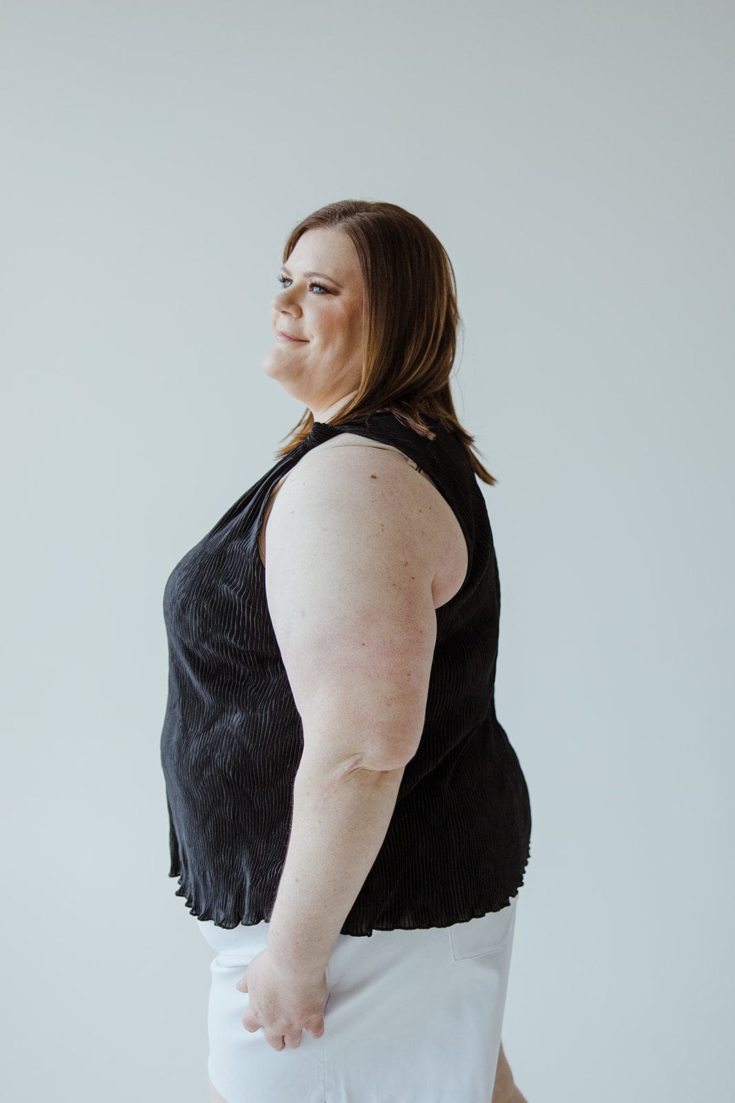 Person with shoulder-length hair wearing a luxurious choice: a sleek Plisse Satin Tank and white shorts, standing against a plain background.