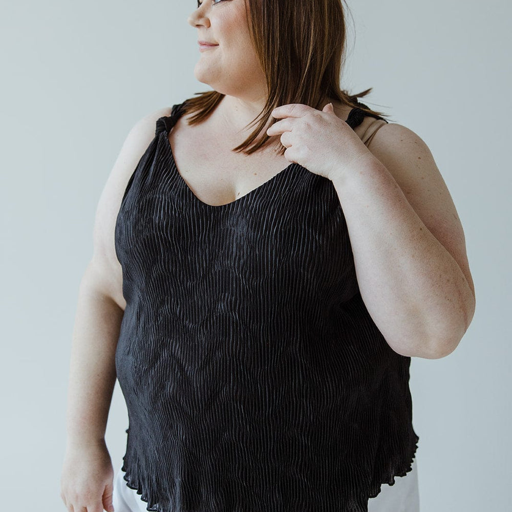 A person with shoulder-length hair wearing a sophisticated Plisse Satin Tank and white shorts looks to the side against a plain background, showcasing a luxurious choice in their black top.