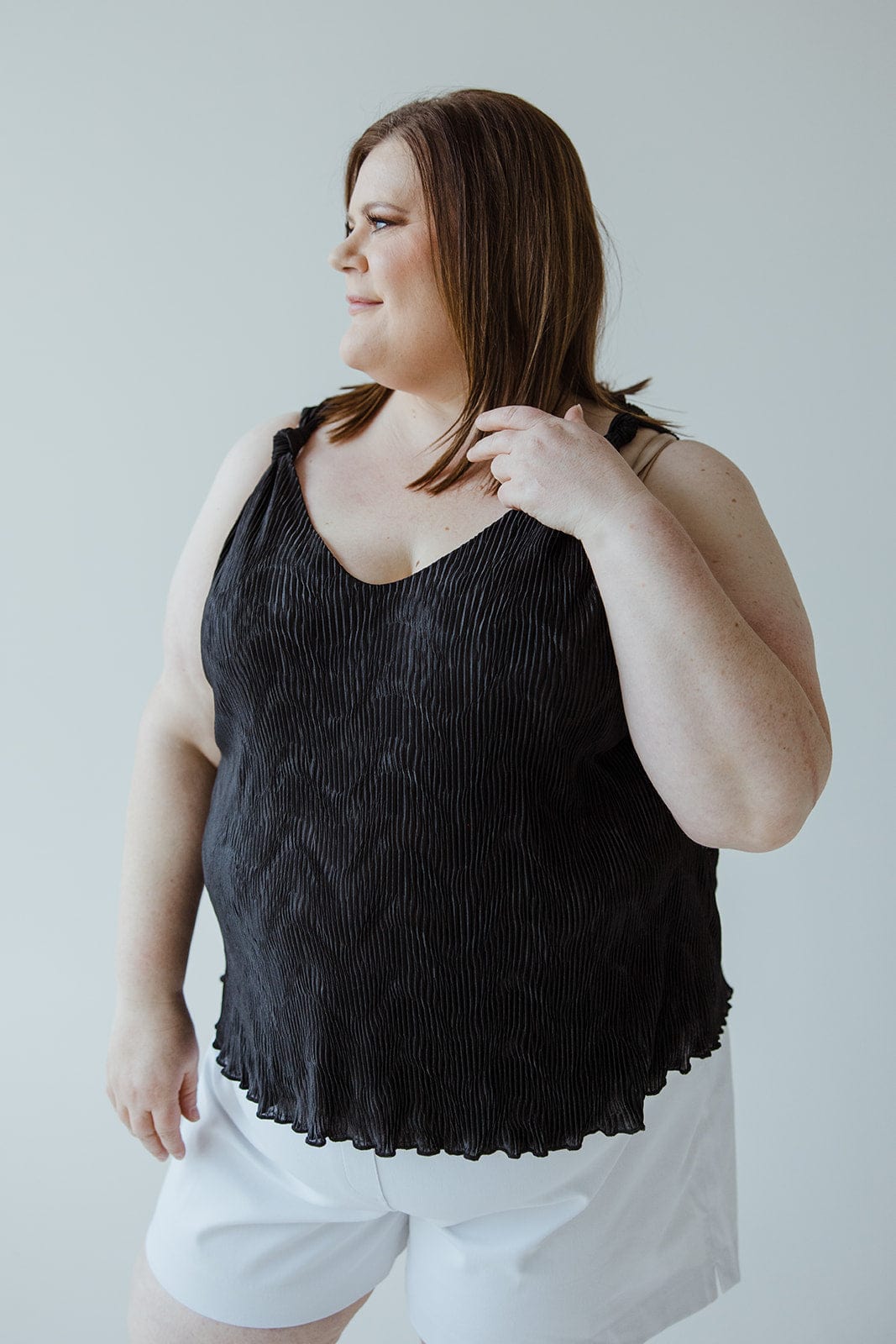 A person with shoulder-length hair wearing a sophisticated Plisse Satin Tank and white shorts looks to the side against a plain background, showcasing a luxurious choice in their black top.