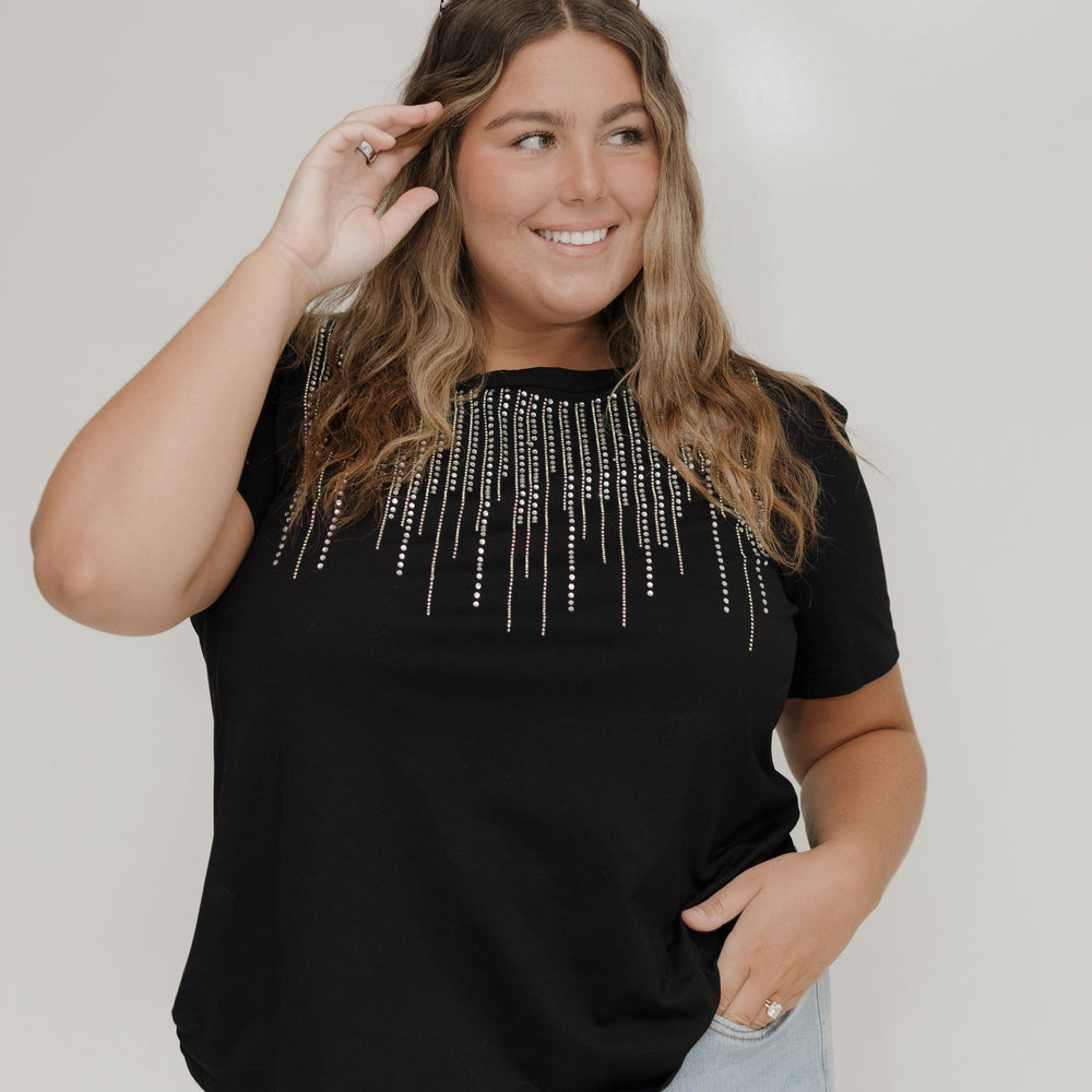 Woman wearing a black top with decorative elements and light blue jeans against a plain background