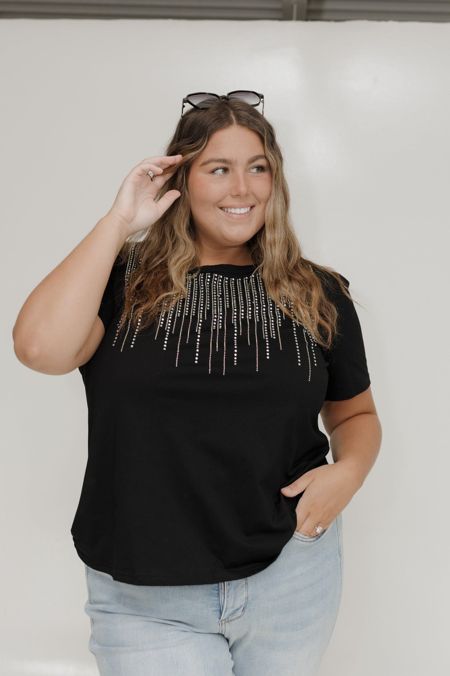 Woman wearing a black top with decorative elements and light blue jeans against a plain background