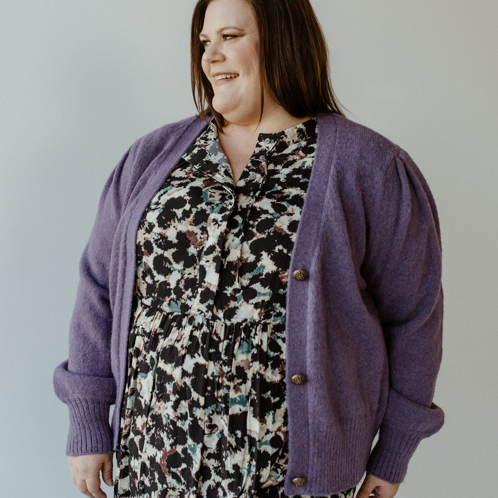 A woman wearing Yesta's PUFF SLEEVE CARDIGAN WITH BUTTONS and a floral dress stands against a plain background, looking to the side with a smile.