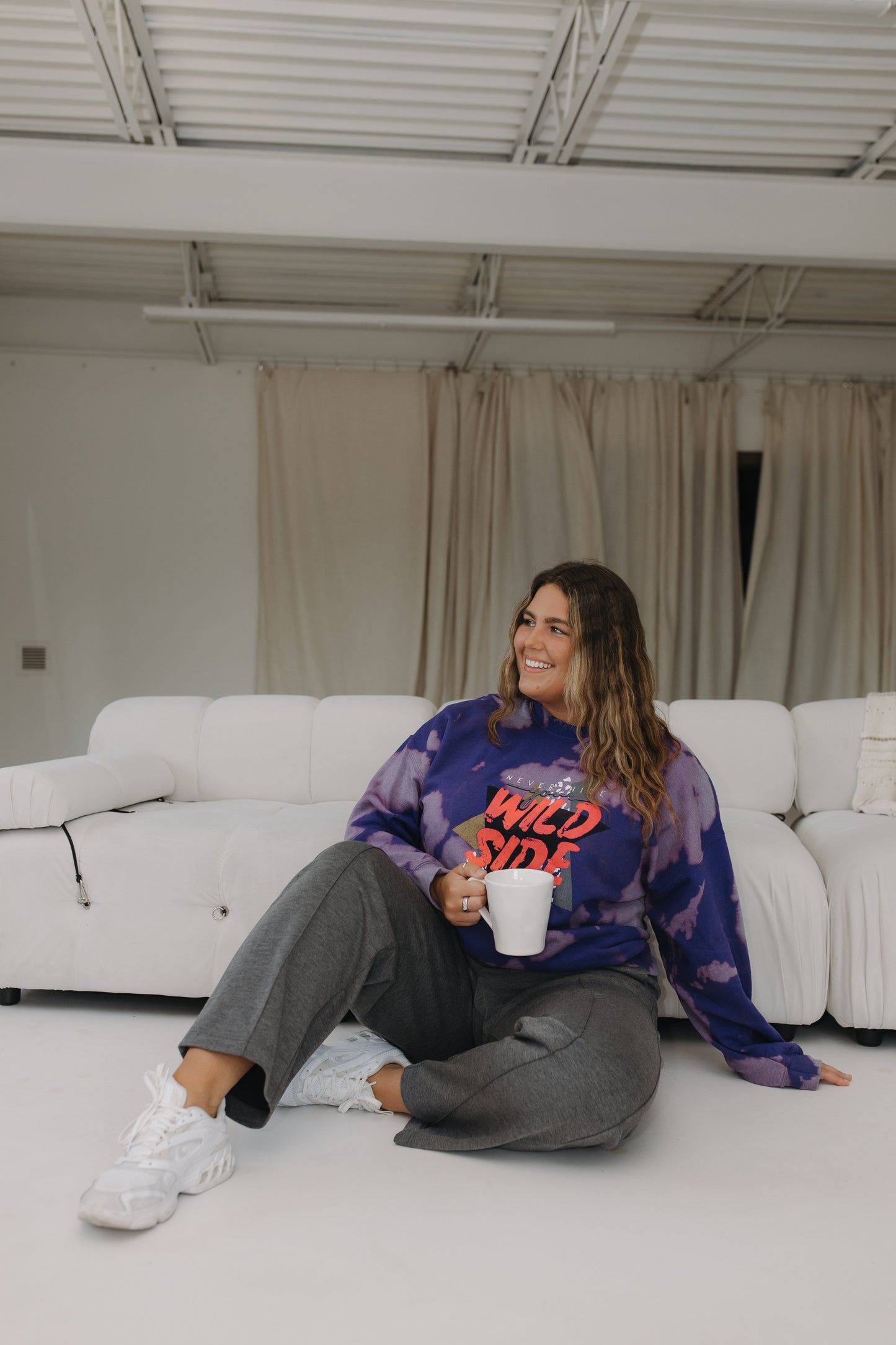Woman sitting on the floor with a white cup, wearing a tie-dye sweatshirt and gray pants, in a room with white walls and a white couch.
