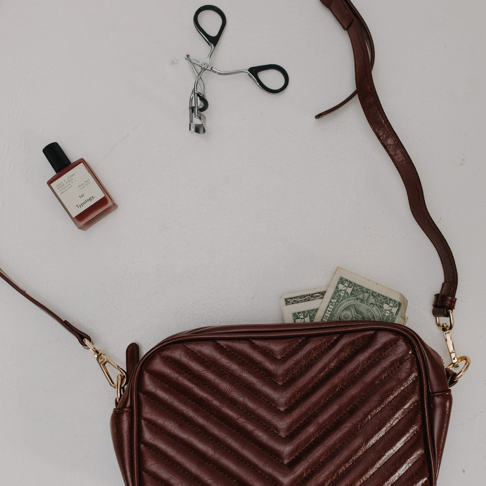 Burgundy chevron-patterned handbag with a strap, small bottle, and money on a light gray background